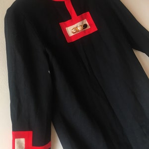 May include: A black dress with red trim and a gold and pearl embellishment on the chest. The dress has long sleeves and a high neckline.
