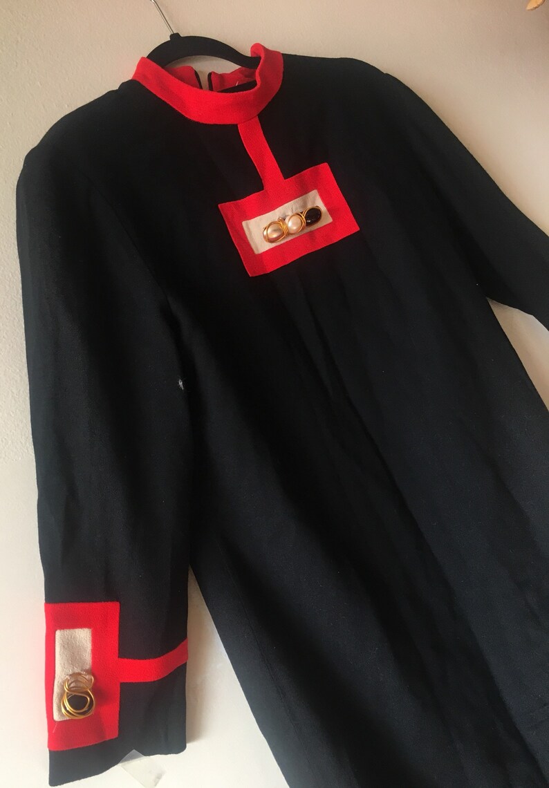May include: A black dress with red trim and a gold and pearl embellishment on the chest. The dress has long sleeves and a high neckline.