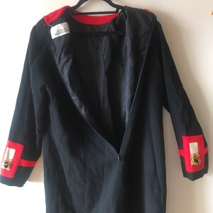 May include: A black long-sleeved dress with red trim around the neckline and cuffs. The cuffs also have a white and gold design. The dress has a zipper down the back. The label inside the dress reads "Steve Fabrikant".