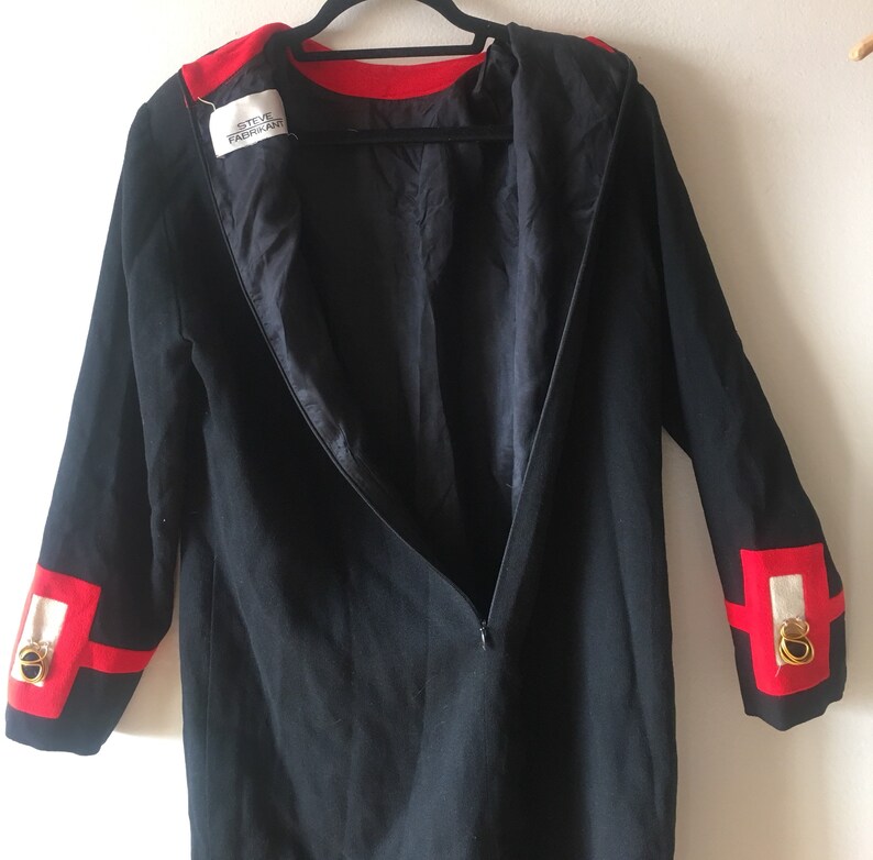 May include: A black long-sleeved dress with red trim around the neckline and cuffs. The cuffs also have a white and gold design. The dress has a zipper down the back. The label inside the dress reads "Steve Fabrikant".