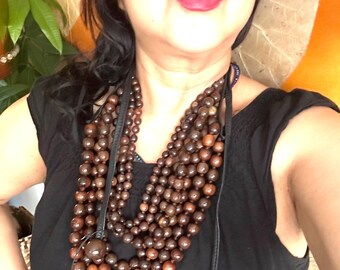 Peut inclure: Un collier à plusieurs rangs avec de grosses perles rondes marron. Le collier a un cordon en cuir et une grosse perle ronde au centre. Les perles varient en teinte du marron clair au foncé. Le collier est porté sur un haut noir.
