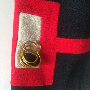 May include: A close-up of a black fabric with red trim. A gold wire design with a black stone is centered in the white fabric square.