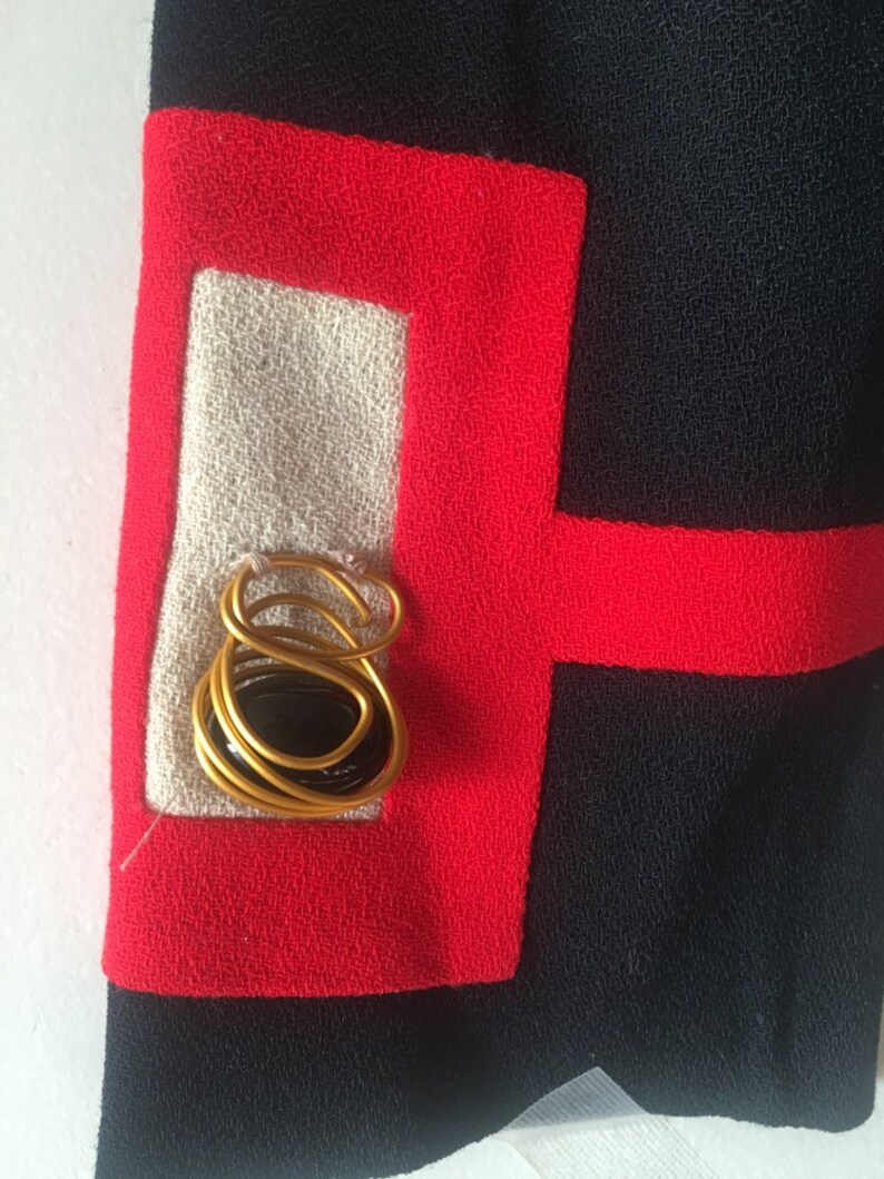 May include: A close-up of a black fabric with red trim. A gold wire design with a black stone is centered in the white fabric square.