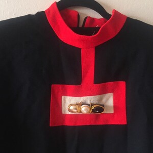 May include: A black long-sleeved sweater with a red trim around the neck and a red square with a white center and gold and black embellishments.