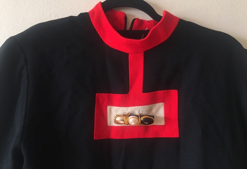 May include: A black long-sleeved sweater with a red trim around the neck and a red square with a white center and gold and black embellishments.