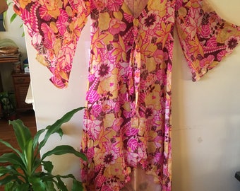 May include: A long, high-low, pink and yellow floral print dress with a V-neck and bell sleeves. The dress has a tie at the waist and is made of a lightweight fabric. The label reads "Sundried Cotton NAMASTE INC."