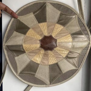 May include: A round, beige leather bag with a patchwork design. The bag has a circular shape and is made of different shades of beige leather, creating a geometric pattern.