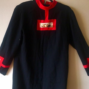 May include: A black long-sleeved dress with red and white trim. The dress has a high neckline and a geometric design on the front and sleeves. The geometric design features gold buttons.