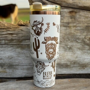 Può includere: Un bicchiere in acciaio inossidabile bianco con un coperchio dorato e un design dorato con mucche delle Highlands, cactus e il testo "Bless Your Heart".