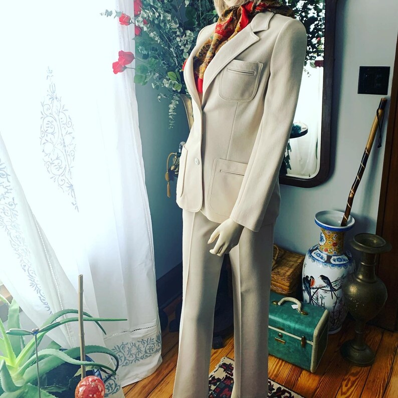 May include: A cream-colored suit on a mannequin. The suit includes a blazer with large pockets and matching pants. A patterned scarf is draped around the mannequin's neck. The suit is displayed in a room with a window and decorative items.