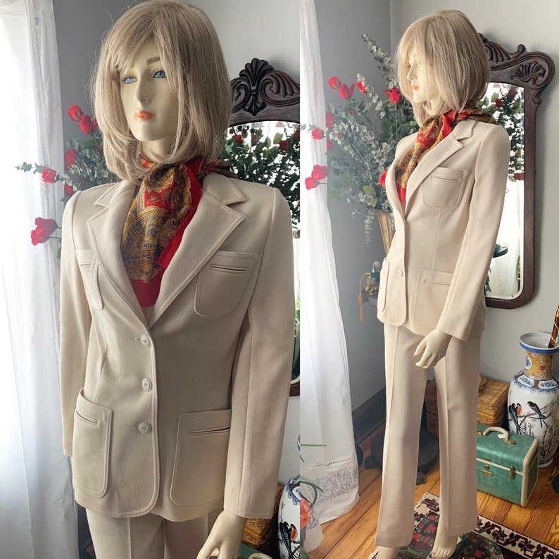May include: A beige two-piece suit with a blazer and matching pants. The blazer has two front pockets and a two-button closure. A red and gold patterned scarf is worn around the neck. The suit is displayed on a mannequin.