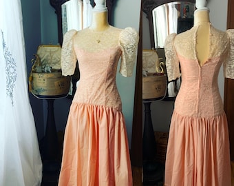 May include: A vintage peach-colored dress displayed on a mannequin. The dress features a lace bodice and sleeves, a high neckline, and a gathered skirt. The back of the dress is also visible, showing a zipper closure. The dress is a classic example of vintage fashion.