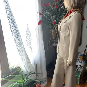May include: A beige pantsuit on a mannequin. The suit includes a blazer with pockets and wide-leg pants. The mannequin is wearing a patterned scarf. The background includes a window with white curtains and a vase of red flowers.