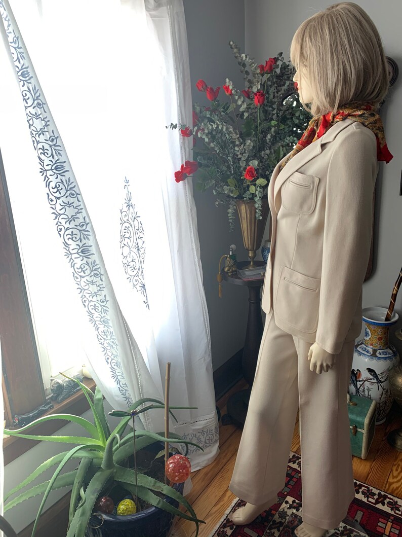 May include: A beige pantsuit on a mannequin. The suit includes a blazer with pockets and wide-leg pants. The mannequin is wearing a patterned scarf. The background includes a window with white curtains and a vase of red flowers.