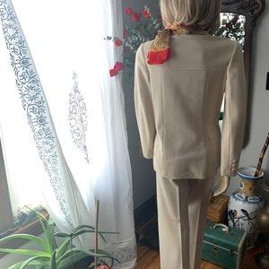 May include: A beige pantsuit with a matching blazer, featuring a classic tailored design. The blazer has three buttons on each cuff. A patterned scarf is draped around the neck. The suit is displayed on a mannequin.