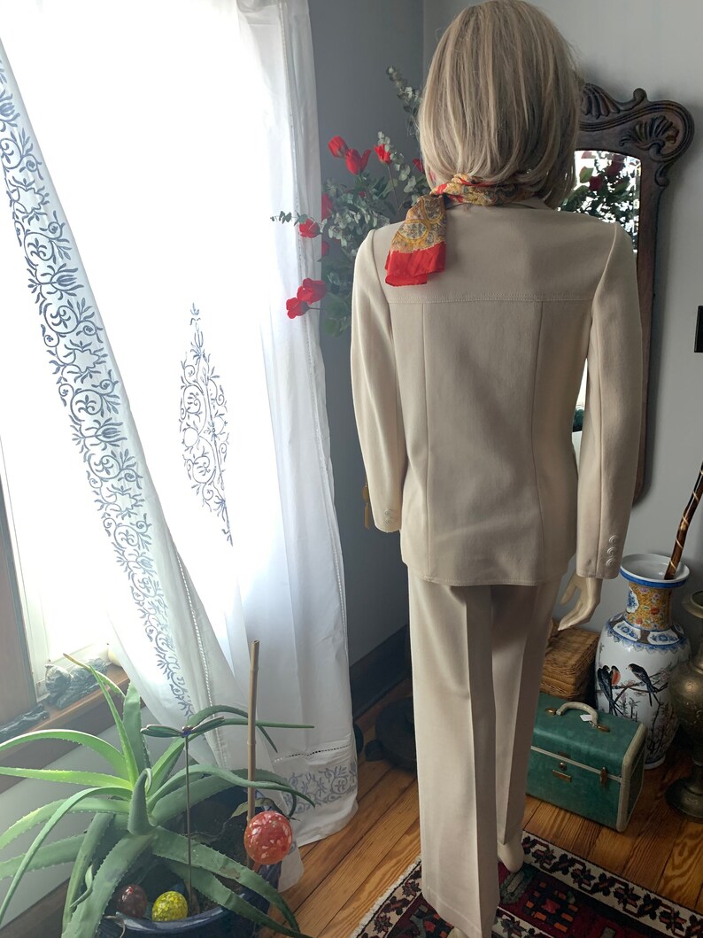 May include: A beige pantsuit with a matching blazer, featuring a classic tailored design. The blazer has three buttons on each cuff. A patterned scarf is draped around the neck. The suit is displayed on a mannequin.