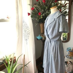 May include: A vintage blue and white gingham dress on a mannequin. The dress has a wide collar and a pleated skirt. The background includes a window with white curtains, a vase of red roses, and a decorative mirror.