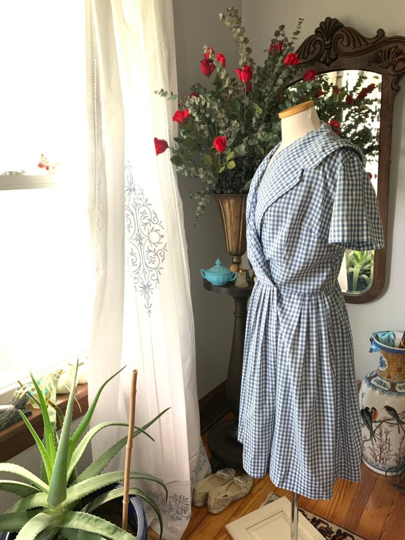 May include: A vintage blue and white gingham dress on a mannequin. The dress has a wide collar and a pleated skirt. The background includes a window with white curtains, a vase of red roses, and a decorative mirror.