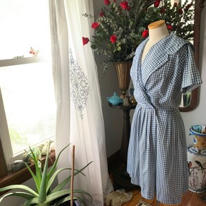 May include: A vintage-style blue and white gingham dress with a wrap-style bodice and a flared skirt. The dress features a wide collar and short sleeves. The dress is displayed on a mannequin in front of a window with white curtains.