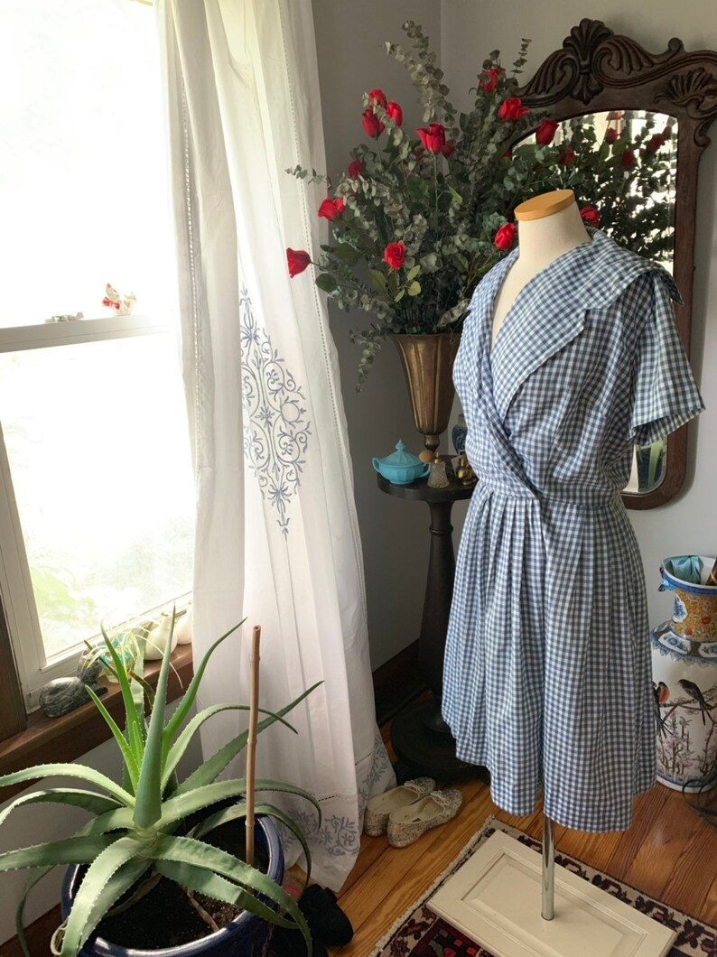 May include: A vintage-style blue and white gingham dress with a wrap-style bodice and a flared skirt. The dress features a wide collar and short sleeves. The dress is displayed on a mannequin in front of a window with white curtains.