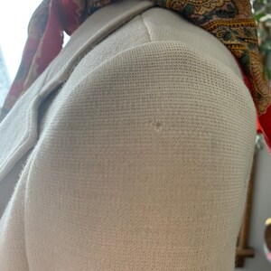 May include: Close-up of a cream-colored blazer with a textured fabric. The blazer's shoulder is visible, along with a portion of the lapel. A patterned scarf in red, green, and gold is draped over the shoulder, adding a pop of color.