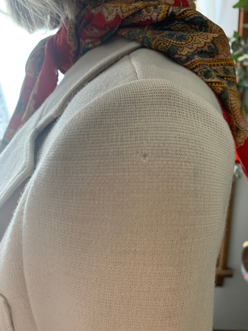 May include: Close-up of a cream-colored blazer with a textured fabric. The blazer's shoulder is visible, along with a portion of the lapel. A patterned scarf in red, green, and gold is draped over the shoulder, adding a pop of color.