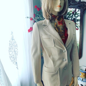 May include: A beige blazer with two front pockets and two button closures. The blazer is worn over a red and gold patterned scarf. The mannequin is displayed in front of a window with white curtains and red roses.