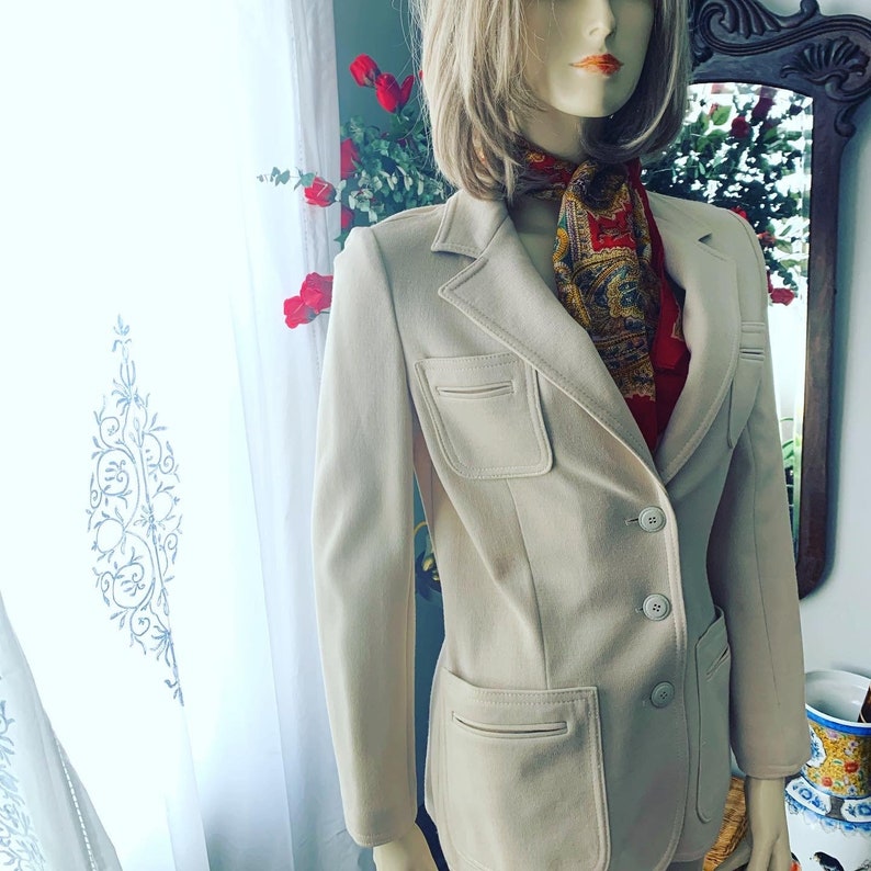 May include: A beige blazer with two front pockets and two button closures. The blazer is worn over a red and gold patterned scarf. The mannequin is displayed in front of a window with white curtains and red roses.