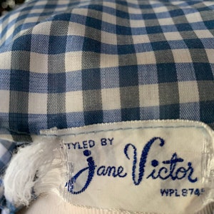 May include: Close-up of a blue and white gingham fabric. The fabric is part of a garment, with a label that reads "STYLED BY Jane Victor". The label is white with blue text and a decorative font.