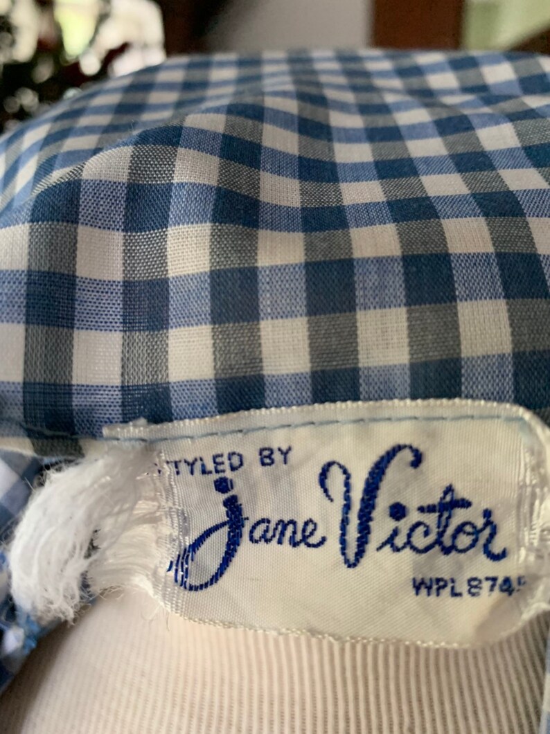 May include: Close-up of a blue and white gingham fabric. The fabric is part of a garment, with a label that reads "STYLED BY Jane Victor". The label is white with blue text and a decorative font.