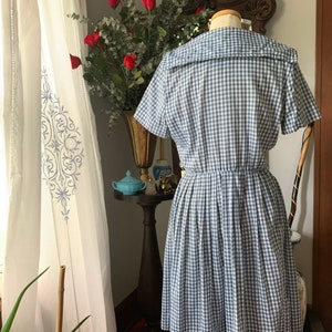 May include: A vintage-style blue and white gingham dress with a sailor collar and short sleeves. The dress has a fitted bodice and a flared skirt. The dress is displayed on a mannequin, showcasing its classic design and silhouette. The dress is a size medium.