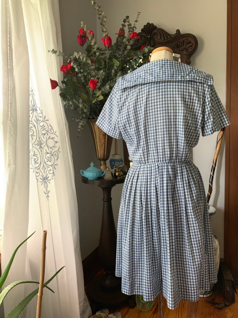 May include: A vintage-style blue and white gingham dress with a sailor collar and short sleeves. The dress has a fitted bodice and a flared skirt. The dress is displayed on a mannequin, showcasing its classic design and silhouette. The dress is a size medium.