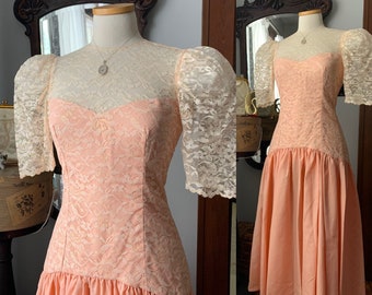 May include: A vintage peach-colored dress with a lace bodice and sheer lace sleeves. The dress features a sweetheart neckline and a gathered skirt. A silver necklace is visible on the mannequin.