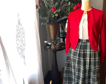 May include: A red cardigan sweater with a white lace blouse underneath, paired with a black and white plaid skirt. The outfit is displayed on a white mannequin against a white wall with a vase of red roses and greenery in the background.
