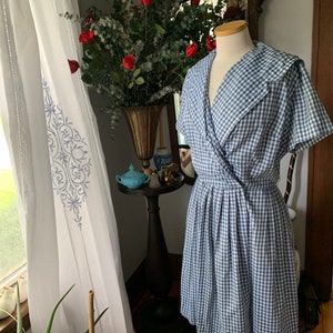 May include: A blue and white gingham dress with a wrap-style bodice and a pleated skirt. The dress is displayed on a mannequin. The dress has a wide collar and short sleeves. The dress is a vintage style.