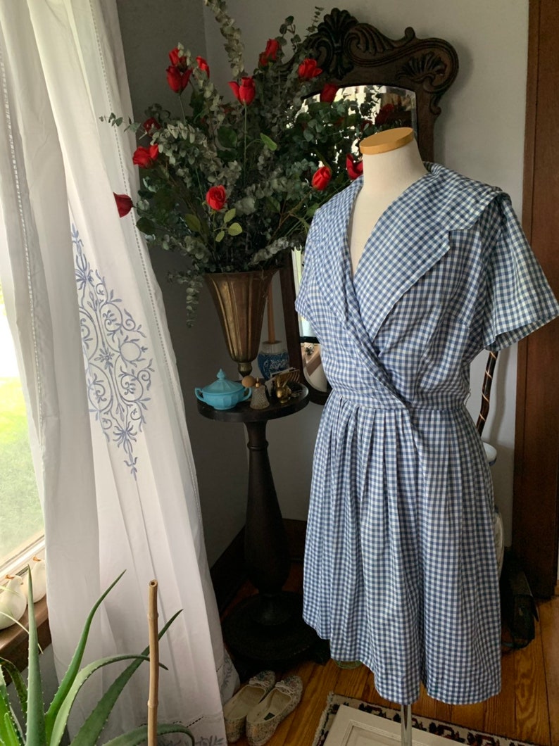 May include: A blue and white gingham dress with a wrap-style bodice and a pleated skirt. The dress is displayed on a mannequin. The dress has a wide collar and short sleeves. The dress is a vintage style.