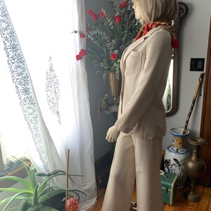 May include: A beige pantsuit on a mannequin. The suit includes a blazer with pockets and matching wide-leg pants. The mannequin is wearing a patterned scarf. The background includes a window with white curtains and a vase of red flowers.