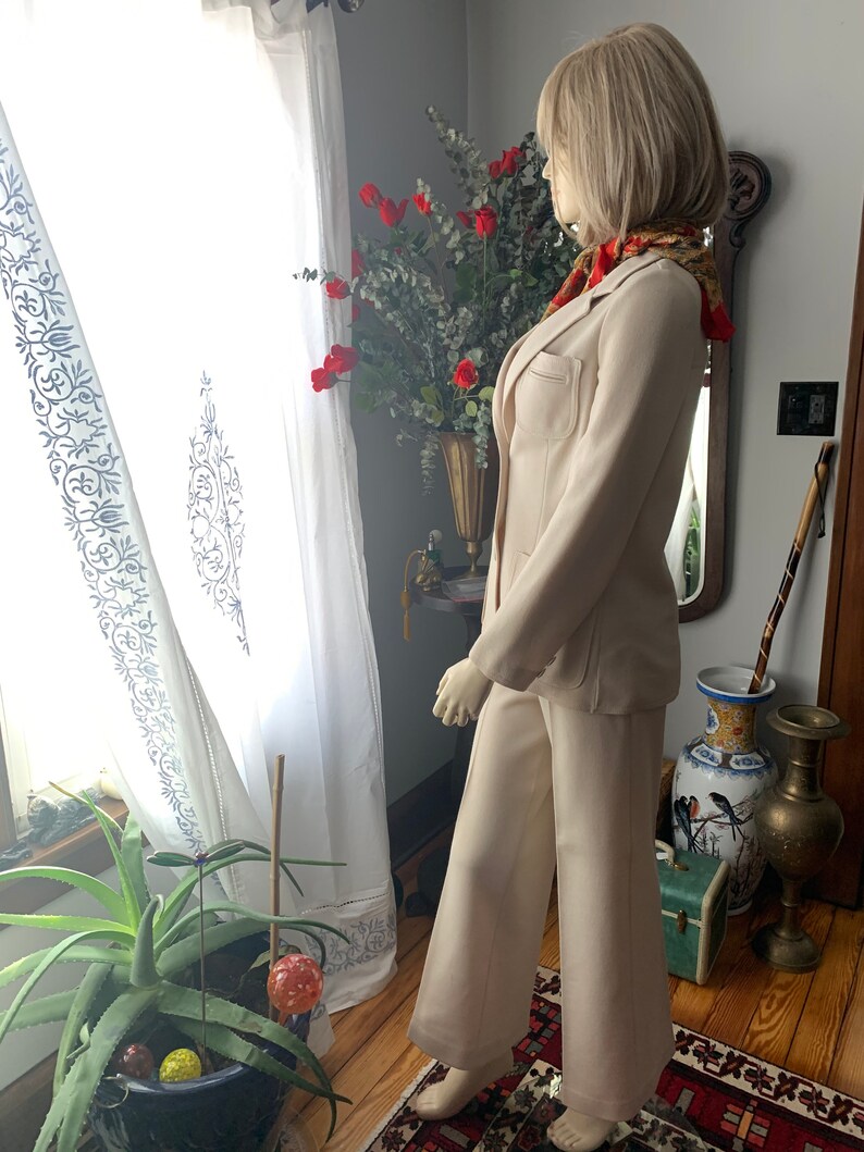 May include: A beige pantsuit on a mannequin. The suit includes a blazer with pockets and matching wide-leg pants. The mannequin is wearing a patterned scarf. The background includes a window with white curtains and a vase of red flowers.