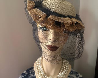 May include: A vintage straw hat with a black ribbon and gold trim, adorned with a black veil. The hat is displayed on a mannequin wearing a pearl necklace.