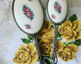 Puede incluir: Un juego de dos espejos de mano vintage con mangos de plata adornados y espejos de forma ovalada. Los espejos tienen un fondo blanco con rosas rosas pintadas a mano y hojas azules.