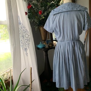 May include: A vintage blue and white gingham dress with a wide collar and short sleeves. The dress is displayed on a mannequin, showcasing its pleated skirt and fitted waist. The dress is a classic style.