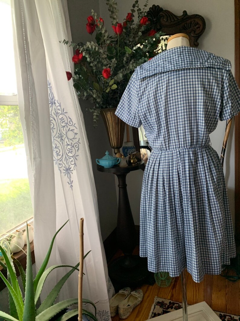 May include: A vintage blue and white gingham dress with a wide collar and short sleeves. The dress is displayed on a mannequin, showcasing its pleated skirt and fitted waist. The dress is a classic style.