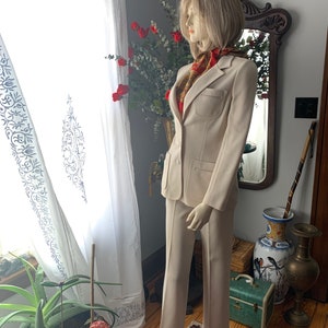 May include: A beige two-piece suit displayed on a mannequin. The suit includes a blazer with a pocket and matching trousers. A patterned scarf adds a pop of color. The mannequin is positioned near a window with white curtains.