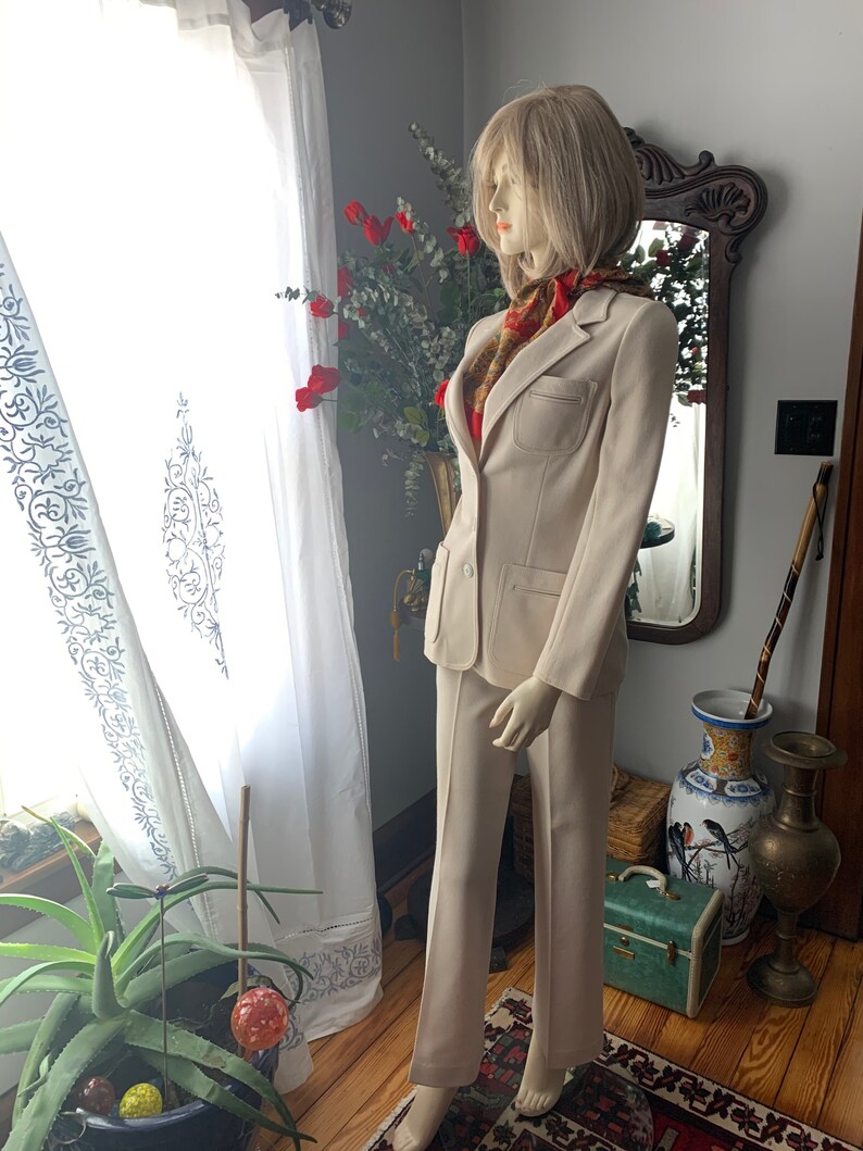May include: A beige two-piece suit displayed on a mannequin. The suit includes a blazer with a pocket and matching trousers. A patterned scarf adds a pop of color. The mannequin is positioned near a window with white curtains.