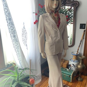 May include: A beige two-piece suit on a mannequin. The suit includes a blazer with two front pockets and matching pants. A patterned scarf is around the mannequin's neck. The mannequin is in front of a window with white curtains.