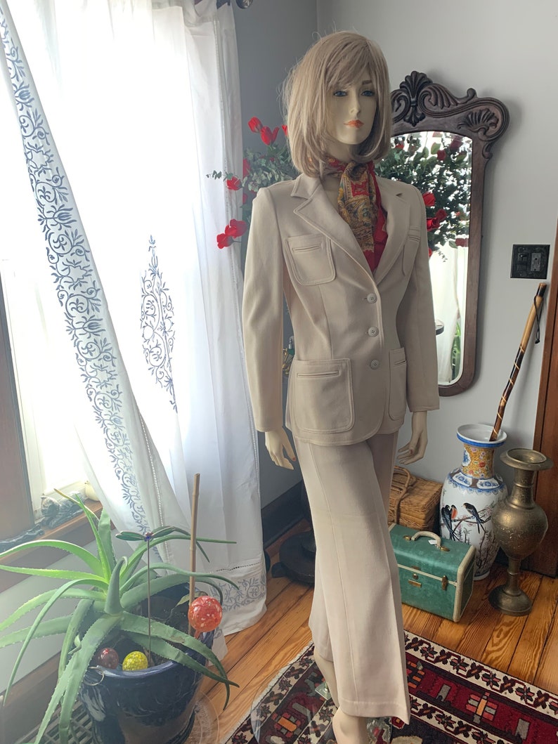 May include: A beige two-piece suit on a mannequin. The suit includes a blazer with two front pockets and matching pants. A patterned scarf is around the mannequin's neck. The mannequin is in front of a window with white curtains.