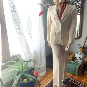 May include: A cream-colored suit displayed on a mannequin. The suit includes a blazer with two front pockets and matching pants. A patterned scarf adds a pop of color. The suit is displayed in a well-lit room with a window and decorative elements.