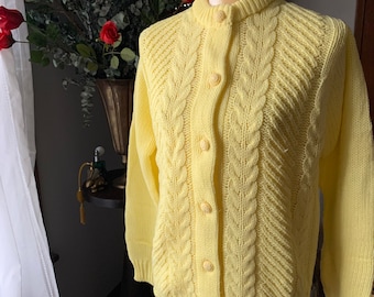 May include: A light yellow cardigan sweater with a cable knit design. The sweater has a round neckline and is buttoned up the front with matching yellow buttons. The sleeves are long, and the sweater is displayed on a mannequin.