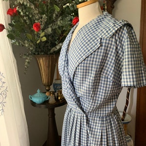 May include: A vintage blue and white gingham dress on a mannequin. The dress features a wrap-style bodice and a pleated skirt. The dress is displayed in front of a vase of red roses and greenery.