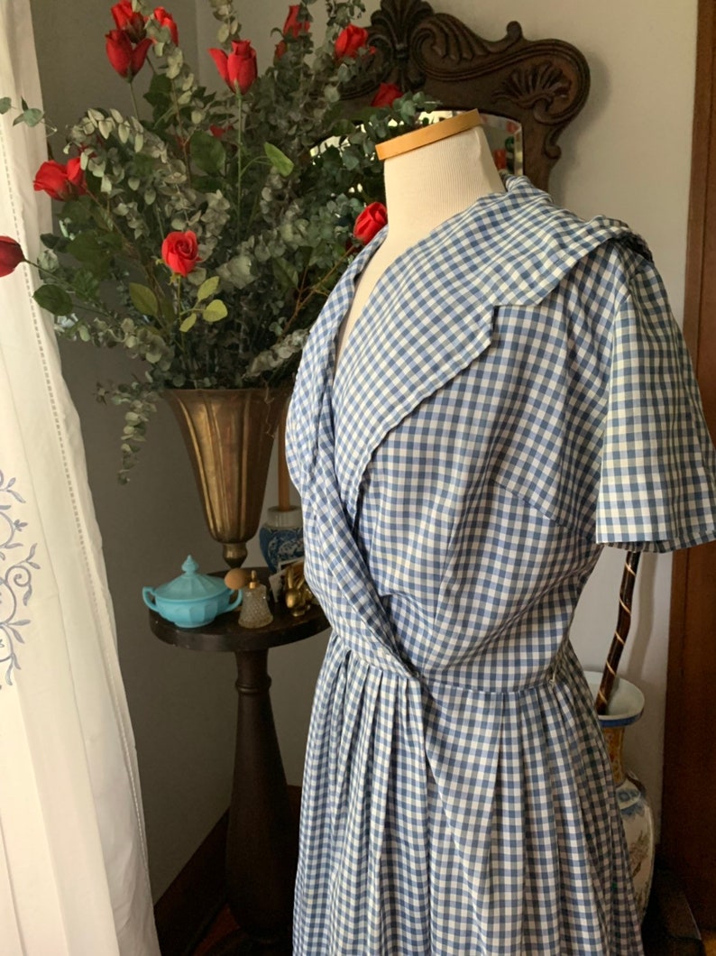May include: A vintage blue and white gingham dress on a mannequin. The dress features a wrap-style bodice and a pleated skirt. The dress is displayed in front of a vase of red roses and greenery.